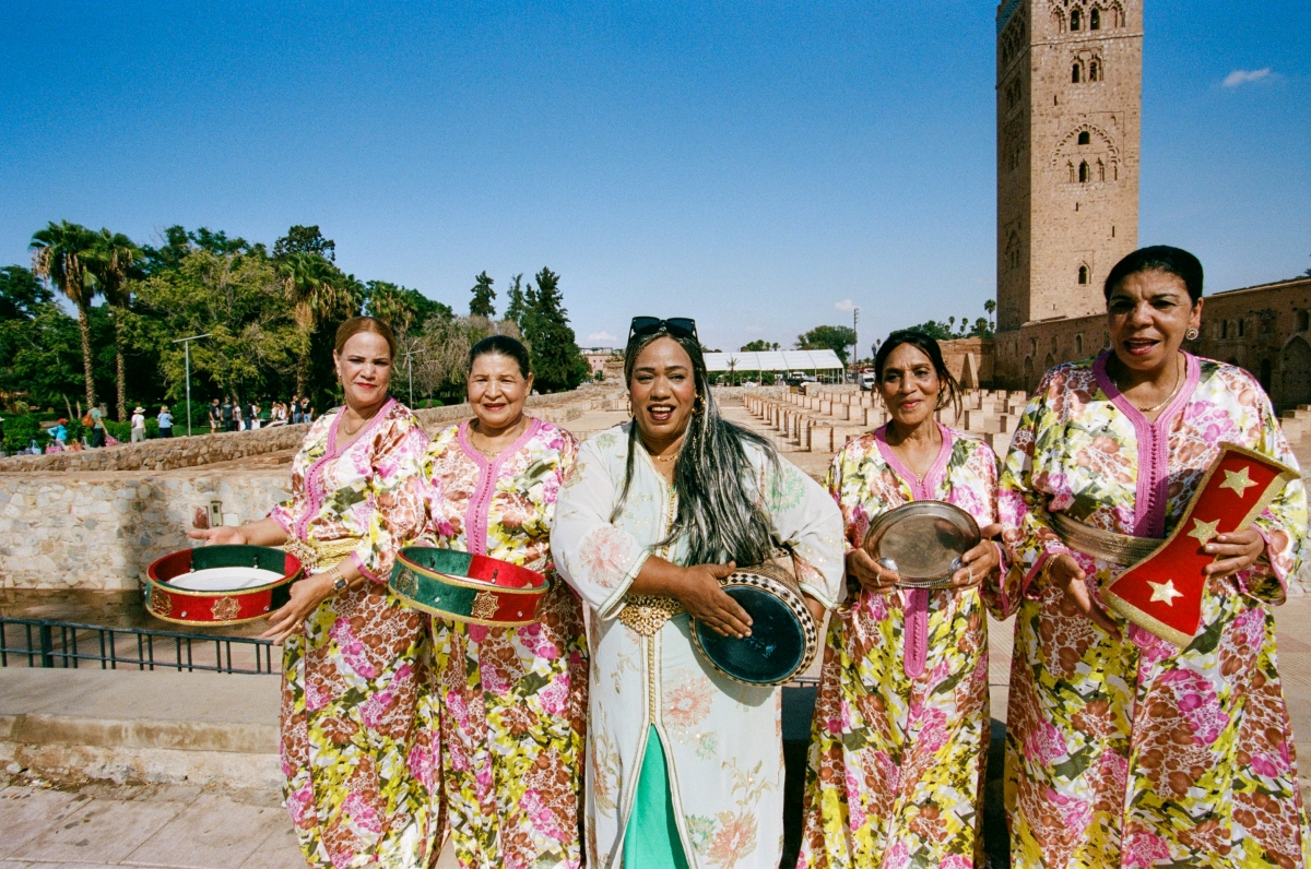 A photo of five women standing in a row. They are all playing a type of drum. The four women on the outside, the Bnat el Houariyat ensemble, are all wearing the same dress with green and pink flowers on it. The woman in the middle, Khadija Warzazia, is wearing a light blue dress.